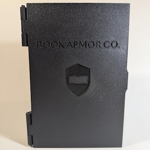 May include: A rectangular black protective book case with a textured finish. The front cover features the text BOOK ARMOR CO. in a serif font above a shield emblem containing a book icon. The case has two visible hinges on the left side.