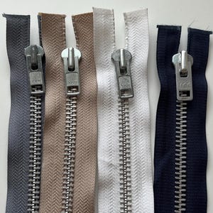 May include: Four separate metal zippers arranged vertically, featuring fabric tapes in dark gray, beige, white, and navy blue. Each zipper includes a silver-toned metal slider and pull tab with the YKK brand name clearly embossed on the metal hardware.