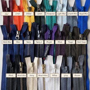 May include: A collection of twenty-eight plastic zippers arranged in three rows, featuring a wide spectrum of colors including red, coral, yellow, green, turquoise, light blue, greyish blue, royal blue, dark blue, blue, navy, purple, plum, taupe, dark taupe, black, metal olive, ivory, metal white, white, grey, and pewter, each labeled with its corresponding color name.