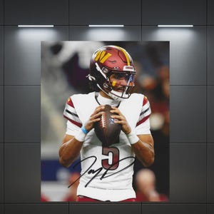 May include: A framed sports photograph featuring a football player in a white jersey with the number 5 and a burgundy and gold helmet, holding a football. The jersey displays a black autograph across the front, capturing a game-day moment.