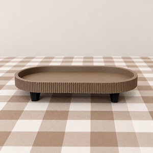 May include: An oval-shaped tray in a muted taupe color featuring a ribbed, textured exterior wall. The tray is elevated by four small, black, block-style feet positioned underneath the base, providing a stable and raised platform for display.