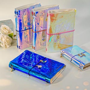 May include: Four iridescent journals with a heart-patterned embossed finish. Each notebook features a colored elastic band closure and small metal charms attached to the spine. The covers reflect light with a holographic sheen.