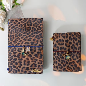May include: Two rectangular notebooks with a brown and black leopard print pattern. The larger notebook features a blue elastic band with two gold-toned charms and the name Sultan in gold script. The smaller notebook has a red elastic band with two gold-toned charms.