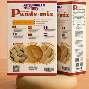 Pode incluir: Duas caixas retangulares de mistura para pão Pinay Co Pande-mix com design em vermelho e branco. A frente exibe texto, listas de ingredientes e imagens circulares de Pandesal, Ensaymada e pão espanhol. O peso líquido é de 530g. Códigos de barras visíveis.