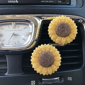 May include: Two gold glitter sunflower car air fresheners hanging from a car's air vent. The air fresheners have brown centers.