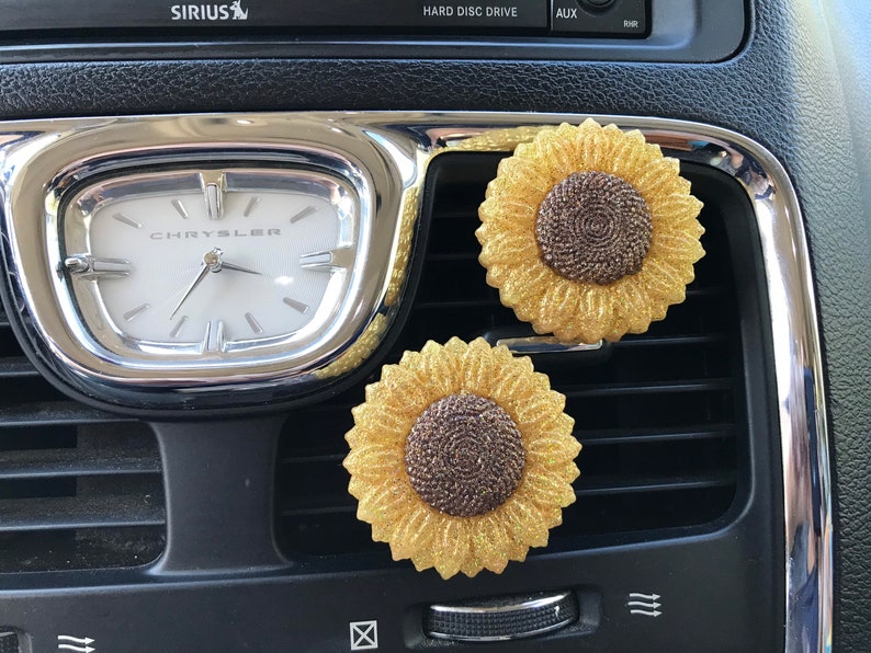 May include: Two gold glitter sunflower car air fresheners hanging from a car's air vent. The air fresheners have brown centers.