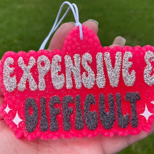 May include: Pink car air freshener with the text "Expensive & Difficult" in silver glitter and black letters.