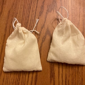 May include: Two white cotton drawstring bags, both closed and lying flat on a wooden surface.