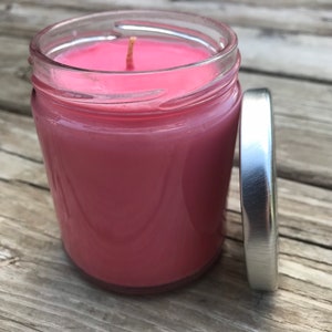 May include: A pink candle in a clear glass jar with a silver lid. The candle is lit and the wick is visible.