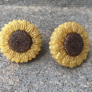 May include: A pair of gold glitter sunflower earrings. The sunflowers have a brown center and are made of resin.