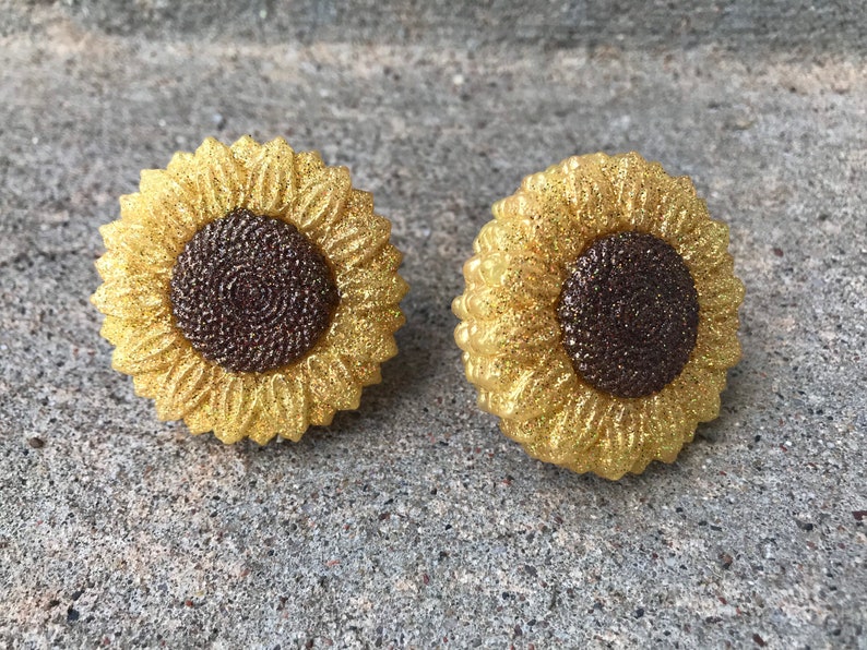 May include: A pair of gold glitter sunflower earrings. The sunflowers have a brown center and are made of resin.