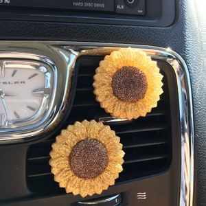 May include: Two gold glitter sunflower car air fresheners. The sunflowers have brown centers and are attached to a car's air vent.