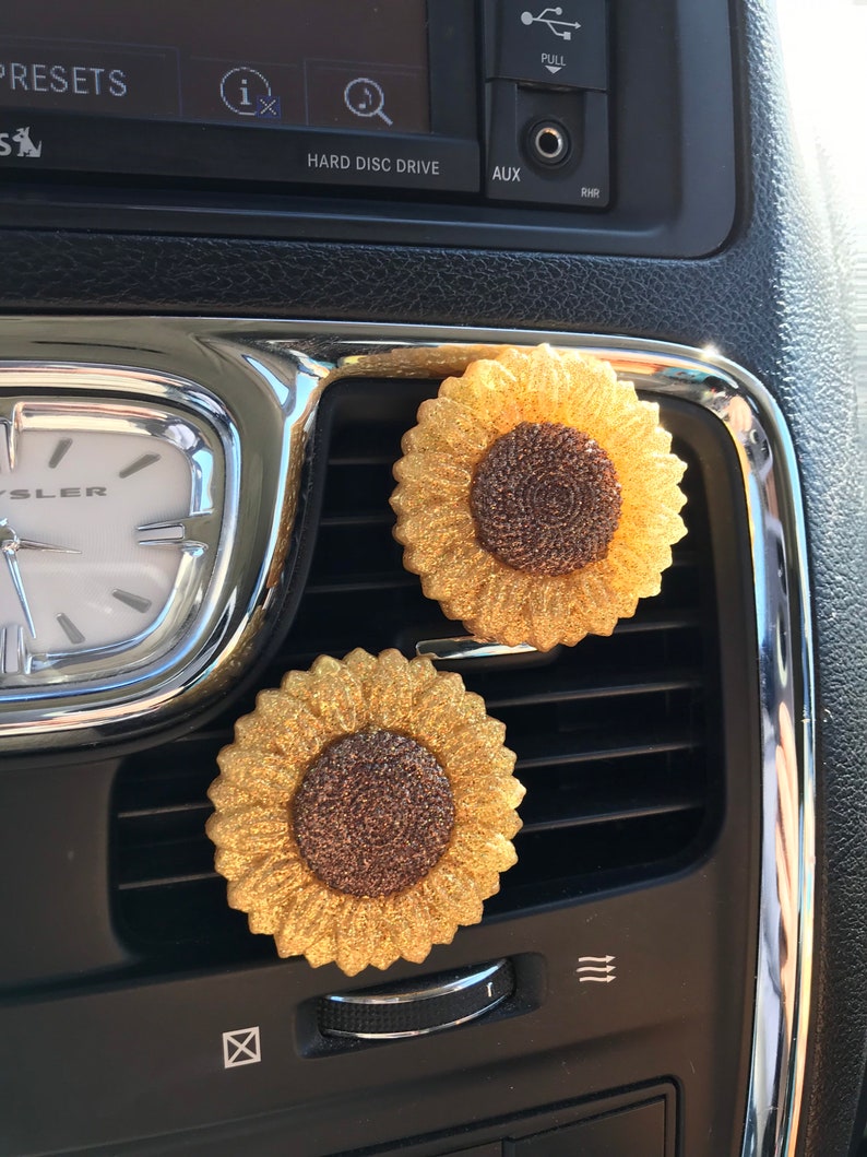 May include: Two gold glitter sunflower car air fresheners. The sunflowers have brown centers and are attached to a car's air vent.