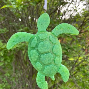 May include: A green sea turtle ornament with a textured surface. The turtle is hanging from a white string.