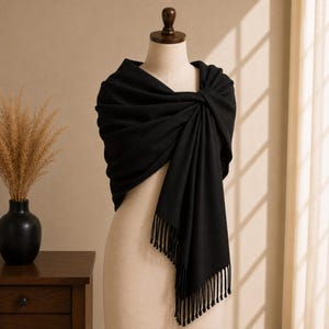 May include: A solid black shawl draped over a mannequin, featuring a soft fabric texture, a gathered knot detail at the shoulder, and long fringe trim along the bottom edge. The wrap is positioned to cover the upper torso with flowing folds.