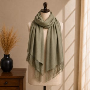 Peut inclure: Une écharpe vert sauge unie drapée sur un mannequin. L'écharpe présente une texture lisse et uniforme et est finie avec de longues franges droites sur les bords inférieurs. Le tissu est enroulé autour du cou dans un pli souple.