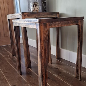 May include: Two nesting tables crafted from dark stained wood with a visible grain pattern. Each table features a square top and four straight legs. The tables are positioned one behind the other, showcasing a matching finish and construction.