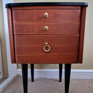 May include: A wooden nightstand with a reddish-brown finish, featuring three drawers with gold-toned hardware. The top surface is black, and the unit is supported by four tapered black legs. The two upper drawers have small knobs, while the bottom drawer has a ring pull.