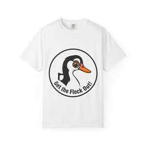 Puede incluir: Una camiseta blanca de manga corta con un gráfico circular en el pecho. El gráfico muestra un pato blanco y negro con el pico y el ojo de color naranja, encerrado en un círculo negro con el texto Get the Flock Out! impreso en negro.