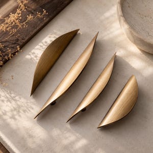 May include: Four curved, crescent-shaped cabinet pulls made of brushed brass metal. Each handle features a smooth, elongated design with pointed ends, showcasing a metallic gold finish with subtle horizontal graining across the surface.