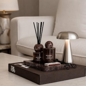 Peut inclure: Un plateau en marbre brun supporte un diffuseur à bâtonnets Saffron Oud avec des tiges noires, une bougie Saffron Oud assortie avec un couvercle sphérique, une petite boîte d'allumettes et une lampe argentée en forme de champignon sur un livre.