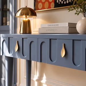 Peut inclure: Une console bleue dotée de deux tiroirs avec des poignées dorées en forme de feuille et un motif de panneaux arqués répétitifs. La surface porte une lampe champignon dorée, deux livres empilés intitulés This Is Home et The Kinfolk Home, et un vase.