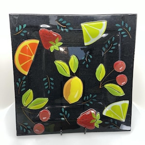 May include: A square black glass plate featuring a colorful fruit motif. The design includes a central yellow lemon, red strawberries, orange slices, lime wedges, and red cherries, all accented with small green leaves and stems.