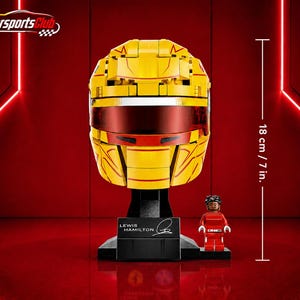 Pode incluir: Um capacete de corrida amarelo construído com blocos, com uma viseira vermelho-escura e detalhes vermelhos. Assenta num suporte preto com uma placa que diz Lewis Hamilton e uma assinatura. Uma pequena figura de fato vermelho mede 18 cm.
