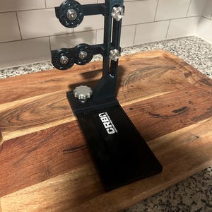 May include: A dark grey fishing rod building stand with a vertical support arm, three adjustable circular tension wheels, and silver star-shaped adjustment knobs. The base features a black plate with the white text CRB CUSTOM ROD BUILDER.