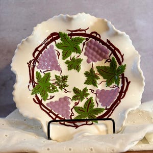 May include: A round, cream-colored ceramic plate with a ruffled edge, featuring a hand-painted design of purple grape clusters, green leaves, and brown vine branches arranged in a circular pattern across the center of the dish.