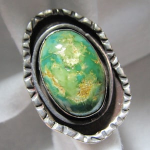 May include: An oval ring featuring a polished green turquoise stone with brown matrix inclusions. The stone is set in a dark, oxidized silver bezel surrounded by a wider, textured silver frame with notched edges and a darkened finish.