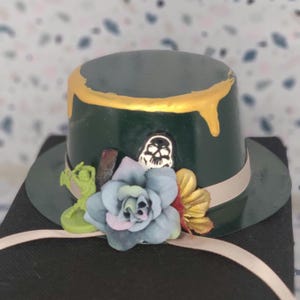 Op de afbeelding: Een donkergroene miniatuur hoge hoed met een goudkleurig druippatroon langs de bovenrand. De hoed is versierd met een lichtblauwe kunstbloem, een klein groen plastic figuurtje, een zwart-wit schedelembleem en een lichtbeige lintband.