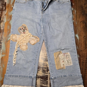May include: Light blue denim capri trousers featuring a large, cream-coloured lace butterfly appliqué on the left leg, a floral and striped fabric patch on the right leg, and white eyelet lace trim along the bottom of both cuffed trouser legs.