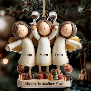 May include: A flat Christmas ornament featuring three stylized figures with brown hair and white robes, each with gold wings. The figures hold wine glasses and stand above small gift boxes. The text reads Lisa, Mary, Emily and Cheers To Another Year.