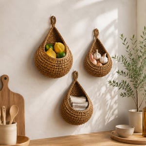May include: Three teardrop-shaped brown woven hanging baskets mounted on a wall. The largest basket holds yellow lemons and green limes, the medium basket contains white garlic bulbs, and the smallest basket stores folded beige and white striped fabric.