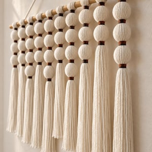 May include: A wall hanging featuring multiple vertical strands of off-white cotton cord suspended from a wooden dowel. Each strand consists of several spherical knots separated by dark brown thread accents, ending in long, loose fringe tassels.
