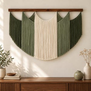 Peut inclure: Une tenture murale composée de longs cordons de coton vert olive et crème suspendus à une barre en bois foncé. Les cordons sont disposés en sections superposées, créant un motif drapé avec une partie centrale claire et des côtés foncés.