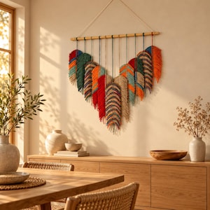 May include: A macrame wall hanging featuring multiple feather-shaped elements suspended from a horizontal wooden rod. The feathers are crafted from multicoloured cotton cord in shades of red, orange, green, blue, and natural beige, arranged in a V-shape.