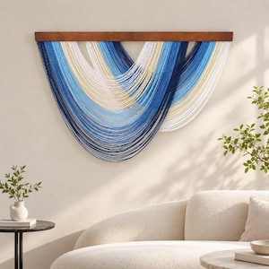 May include: A wall hanging featuring a horizontal dark wood bar with multiple strands of yarn in shades of navy blue, light blue, white, and cream. The yarn is arranged in overlapping, U-shaped swags that create a layered, cascading effect.