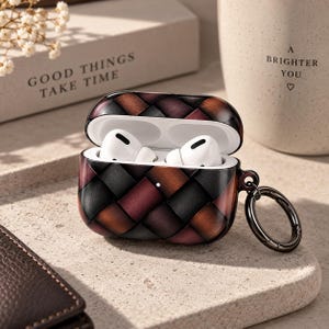 May include: A protective case for wireless earbuds featuring a woven leather-style pattern in shades of black, brown, and deep red. The case is open, revealing white earbuds inside, and includes a dark metal circular carabiner clip attached to the side.