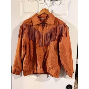 May include: A rust-colored jacket featuring a full-length front zipper, a pointed collar, and long sleeves with ribbed cuffs. The garment is accented with a horizontal fringe detail across the chest and back, extending from the shoulders to the center.