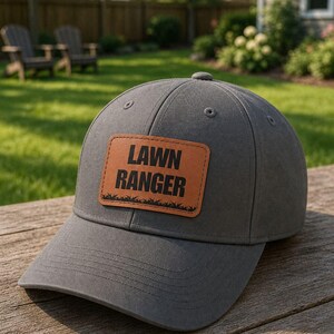 Könnte beinhalten: Eine anthrazitfarbene Baseballkappe mit einem rechteckigen, hellbraunen Aufnäher in Lederoptik auf der Vorderseite. Der Aufnäher zeigt den schwarzen Schriftzug LAWN RANGER über einer Grasgrafik. Die Kappe hat eine strukturierte Krone und einen gebogenen Schirm.