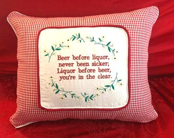 May include: Red and white gingham pillow with a white embroidered square featuring a green wreath with white flowers and the text "Beer before liquor, never been sicker; Liquor before beer, you're in the clear."