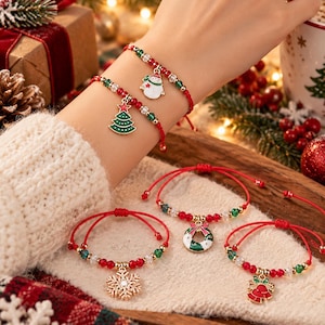 Peut inclure: Un ensemble de bracelets en cordon rouge avec des breloques festives représentant un bonhomme de neige, un sapin de Noël, une couronne, un flocon de neige et une cloche. Chaque bracelet est orné de petites perles rouges, vertes et transparentes.