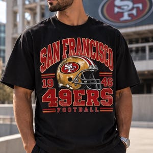 Könnte beinhalten: Ein schwarzes Kurzarm-T-Shirt mit einem Grafikdruck eines goldenen Football-Helms und dem Text SAN FRANCISCO, 19 46, 49ERS sowie FOOTBALL in roter und weißer Schrift, ergänzt durch horizontale rote und gelbe Akzentlinien im Design.