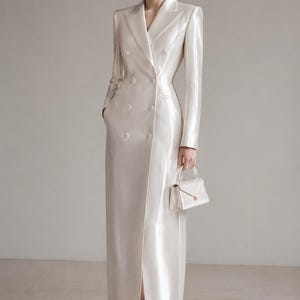 Puede incluir: Un vestido abrigo largo de doble botonadura en tela color blanco roto brillante con cuello estructurado, mangas largas y una abertura frontal. La prenda cuenta con seis botones y se combina con un pequeño bolso de mano estructurado a juego.