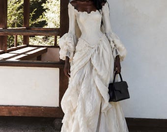 May include: An off-white floor-length gown with a corset-style bodice, long sleeves featuring voluminous ruffled cuffs, and a textured, layered skirt with a scalloped neckline. A small, structured black handbag is held in one hand.