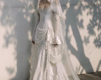 May include: A floor-length white gown with a corset-style bodice, long sheer sleeves, and a draped skirt featuring ruffled accents. The ensemble includes a long lace-trimmed veil and a small white crescent-shaped handbag with a hanging tassel.