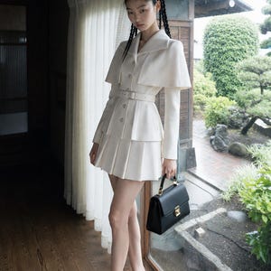 May include: A cream-colored blazer with a cape-style overlay, a matching pleated mini skirt, and a thin belt at the waist. The outfit is paired with black pointed-toe high heels and a structured black handbag with a gold-tone metal clasp.