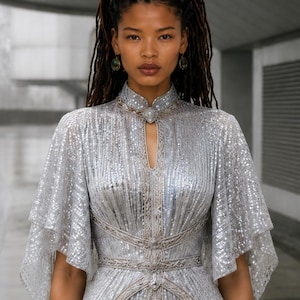 May include: A silver sequined dress featuring a high mandarin collar with a central keyhole cutout, wide flutter sleeves, and intricate silver embroidery detailing along the waistline and neckline, creating a structured and shimmering appearance.